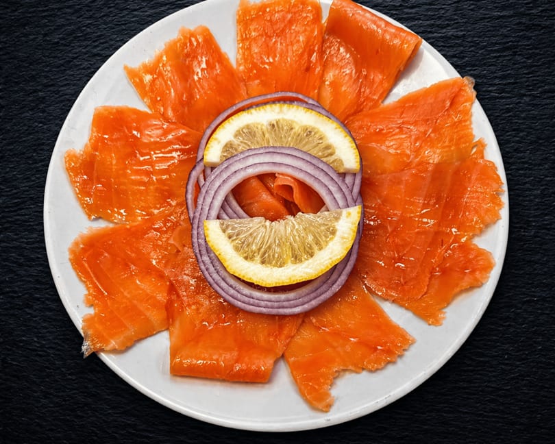 Smoked salmon slices arranged on a white plate with red onion rings and lemon wedges.