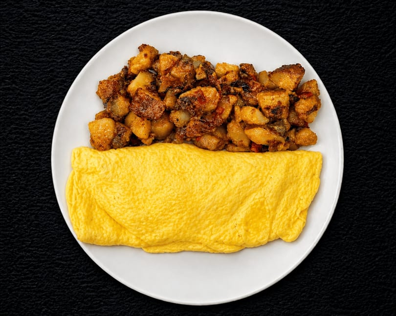 A white plate holds a fluffy yellow omelet and a serving of seasoned breakfast potatoes.