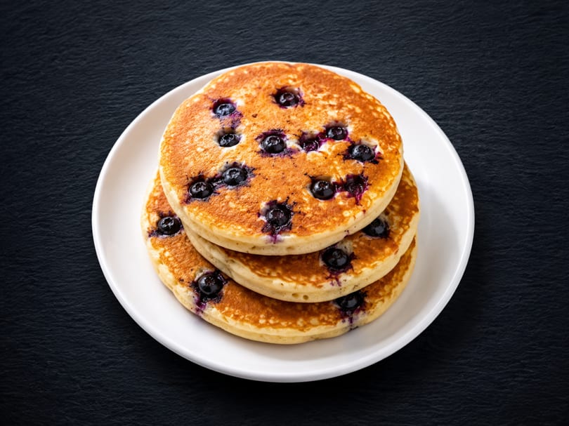 Stack of three golden blueberry pancakes on a white plate against a dark background.