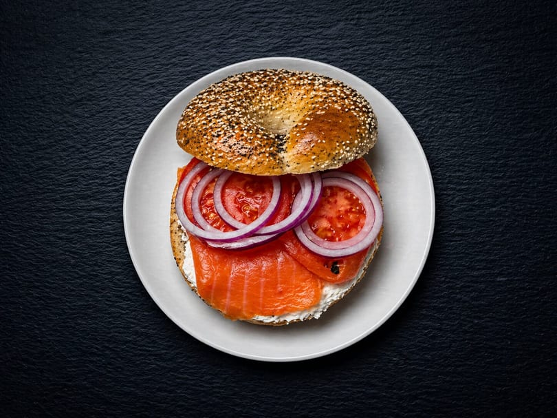 Everything bagel with cream cheese, smoked salmon, tomato, and red onion on a white plate.