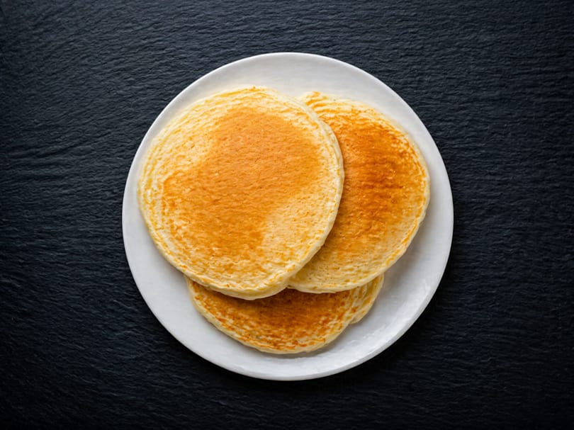 Three golden-brown pancakes stacked on a white plate, centered on a dark, textured surface.