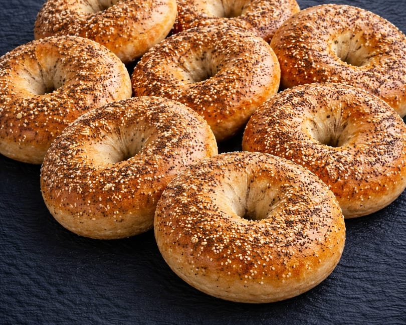 Several golden-brown everything bagels, generously topped with seeds, arranged on a dark surface.